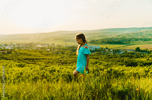 Portrait of little caucasian girl in blue dress walking on a hill at sunset backlit by golden sun light