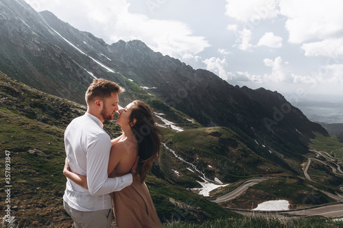 Aerial view of romantic wedding couple of groom and bride in gorgeous wedding dress with long loop standing on the rocks in the green mountains