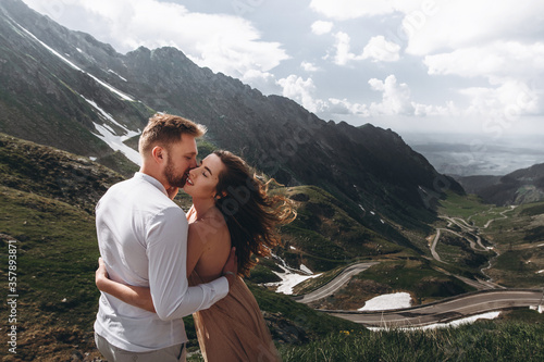 Aerial view of romantic wedding couple of groom and bride in gorgeous wedding dress with long loop standing on the rocks in the green mountains