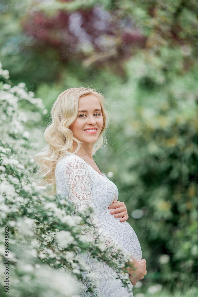 Naklejka premium Young beautiful pregnant woman in a lace white dress walks in a flowering park. Portrait of a beautiful pregnant blonde. Spring.