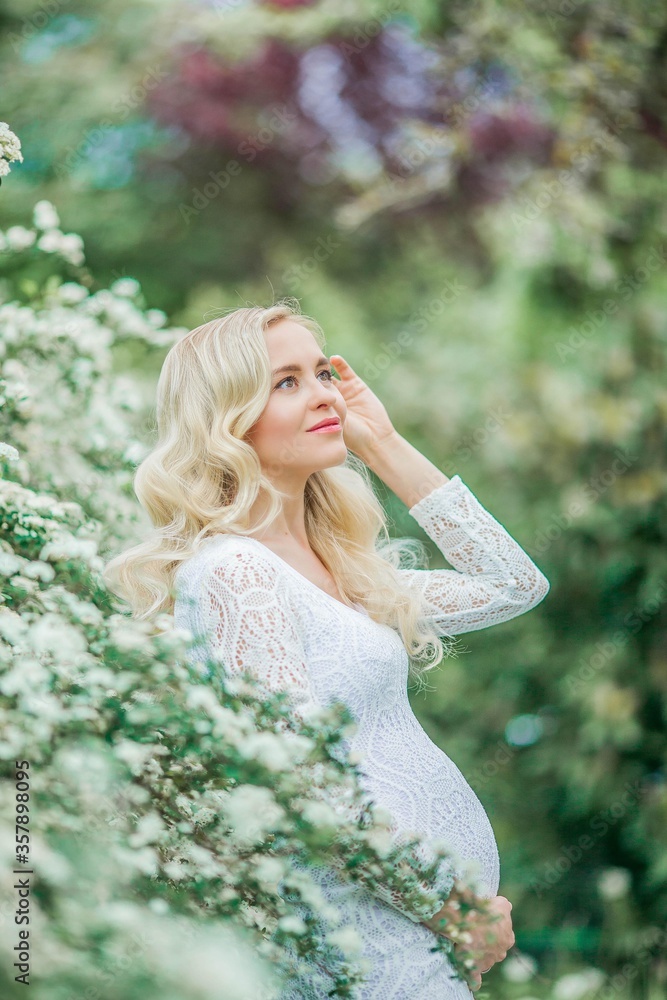 Young beautiful pregnant woman in a lace white dress walks in a flowering park. Portrait of a beautiful pregnant blonde. Spring. Young beautiful pregnant woman in a lace white dress walks in a flowering park. Portrait of a beautiful pregnant blonde. Spring.