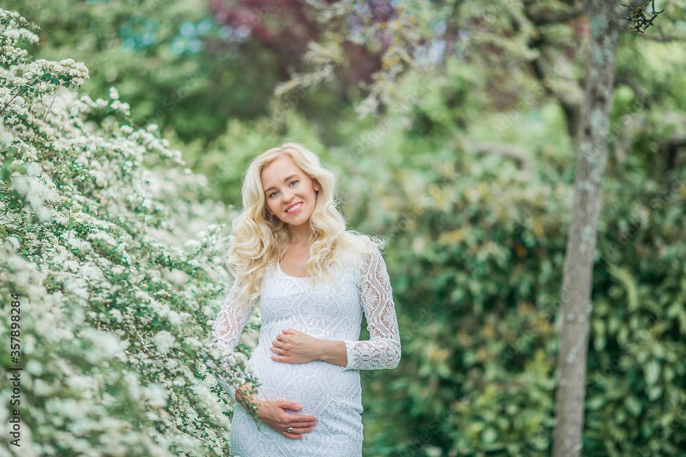 Young beautiful pregnant woman in a lace white dress walks in a flowering park. Portrait of a beautiful pregnant blonde. Spring. Young beautiful pregnant woman in a lace white dress walks in a flowering park. Portrait of a beautiful pregnant blonde. Spring.