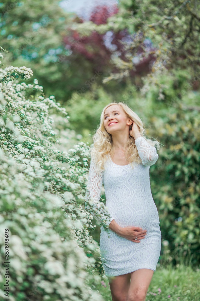 Young beautiful pregnant woman in a lace white dress walks in a flowering park. Portrait of a beautiful pregnant blonde. Spring. Young beautiful pregnant woman in a lace white dress walks in a flowering park. Portrait of a beautiful pregnant blonde. Spring.