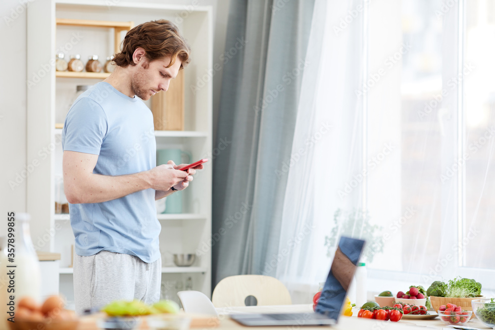 Young man typing a message on mobile phone while standing in the kitchen in the morning Young man typing a message on mobile phone while standing in the kitchen in the morning