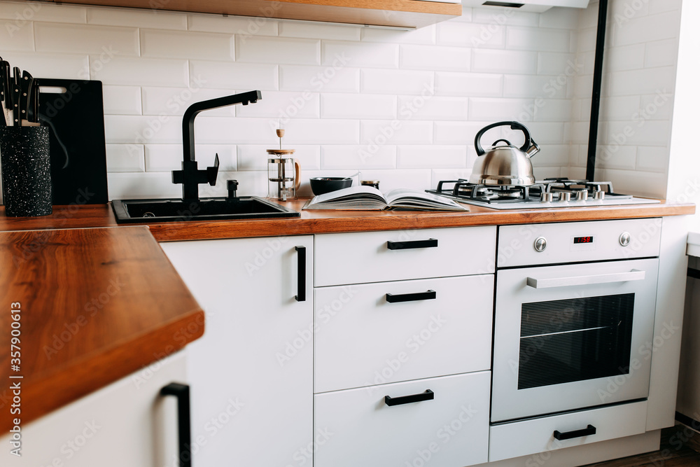 Fototapeta premium Big cook book on the wooden table. Metallic teapot on the gas oven. White kitchen interior. Wooden complete kitchen with gas oven.