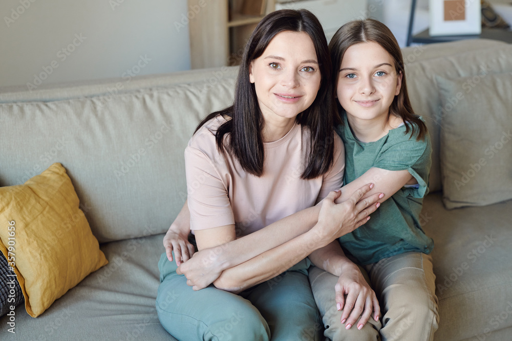 Portrait of content brunette mother sitting on sofa and hugging teenage daughter at home