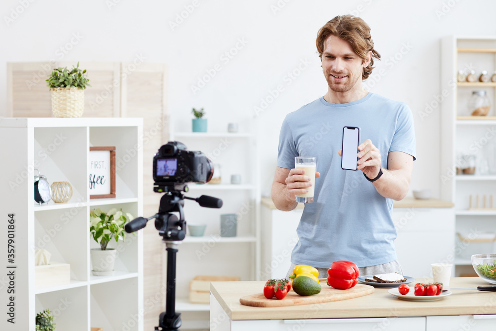Young man using his mobile phone and preparing healthy cocktail from vegetables and shooting the blog on camera