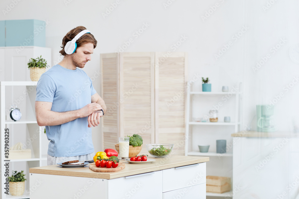Man in headphones checking the time on his watch while standing in domestic kitchen he is going to make dinner Man in headphones checking the time on his watch while standing in domestic kitchen he is going to make dinner