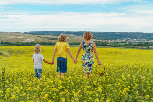 Back view of a beautiful family, a mother and two sons on a yellow flowering field of rapeseed, holding hands. Walking in the open air in nature. Mother in a dress, sons in t-shirts and shorts.