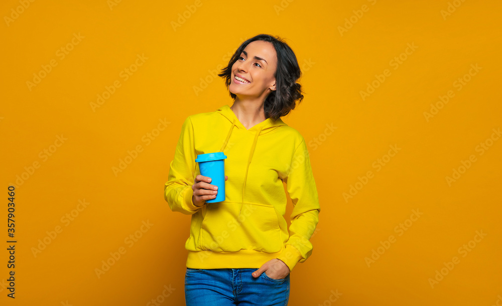 Looking confidently to the future. Half-length photo of smiley girl, confidently looking up and sideways, holding a blue mug in her right hand, dressed in a yellow hoodie. Looking confidently to the future. Half-length photo of smiley girl, confidently looking up and sideways, holding a blue mug in her right hand, dressed in a yellow hoodie.