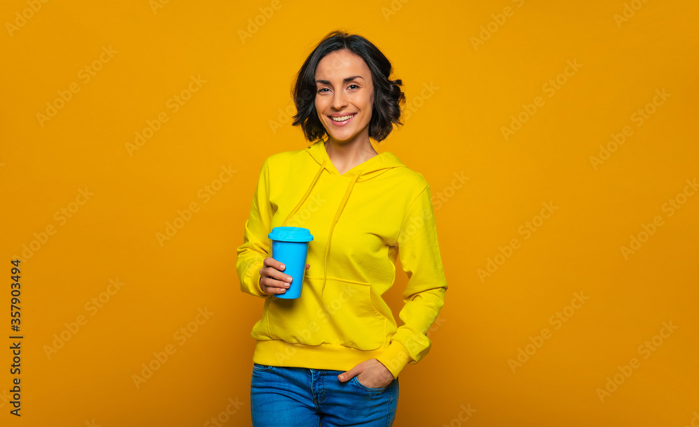 Relaxed day. Half-length photo of smiley relaxed girl dressed in a yellow hoodie, with her left hand in a pocket, looking straight, holding a blue mug in her right hand. Relaxed day. Half-length photo of smiley relaxed girl dressed in a yellow hoodie, with her left hand in a pocket, looking straight, holding a blue mug in her right hand.
