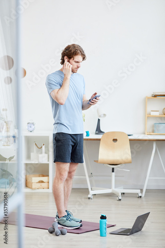 Young man standing on exercise mat and using his mobile phone during sports training at home