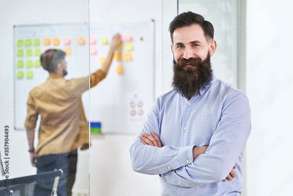 Obraz premium Happy smiling bearded senior developer or manager in modern IT office. Another professional working with scrum desk on background. Successful technology project or startup concept. High quality image.