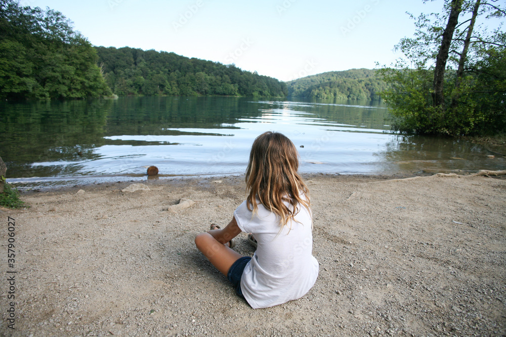 Back view of a blonde child sitting on the beach watching the lake in Plitvice National Park in Croatia.