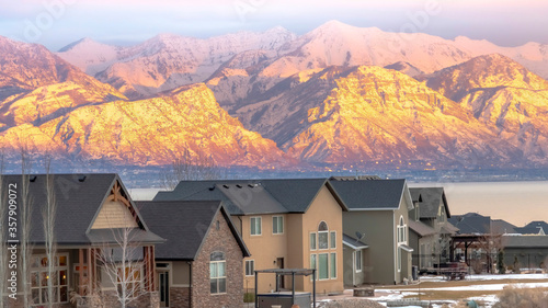 Panorama frame Facade of homes with snowy sunlit mountain and calm lake background at sunset © Jason