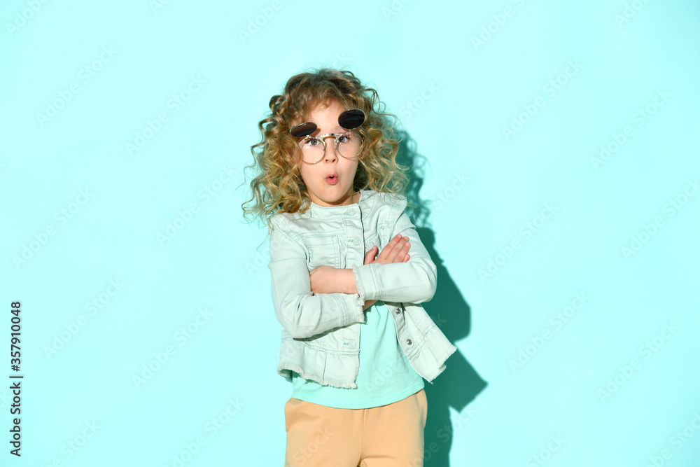 Lovely blond girl with curly hair posing in stylish flip-up glasses calling someone holding hands crossed on chest, isolated on turquoise Lovely blond girl with curly hair posing in stylish flip-up glasses calling someone holding hands crossed on chest, isolated on turquoise