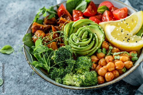 Buddha bowl salad with baked sweet potatoes, chickpeas, broccoli, tomatoes, greens, avocado, pea sprouts on light blue background with napkin. Healthy vegan food, clean eating, dieting, closeup