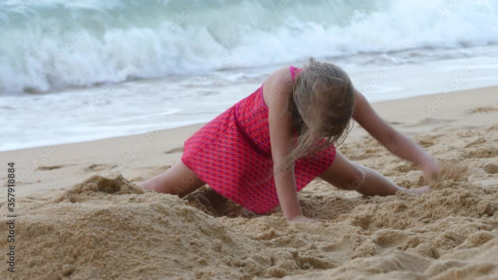 Child girl in red swimsuit dig and play in sand on seashore. Splash ...