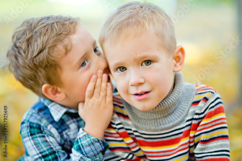 A boy tells a secret to his friend in the ear in an autumn Park against a background of yellow leaves. The boy is surprised by what he has heard.