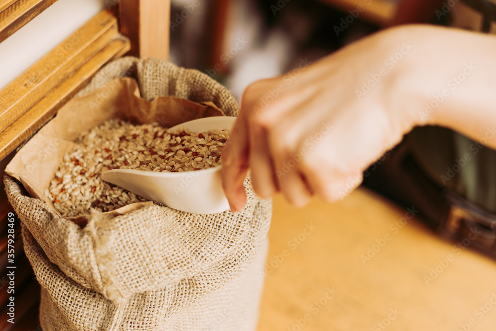 Woman measuring brown rice dosage, taking one plastic scoop of this healthy cereal out of jute clouth bag. Shopping in bulk.