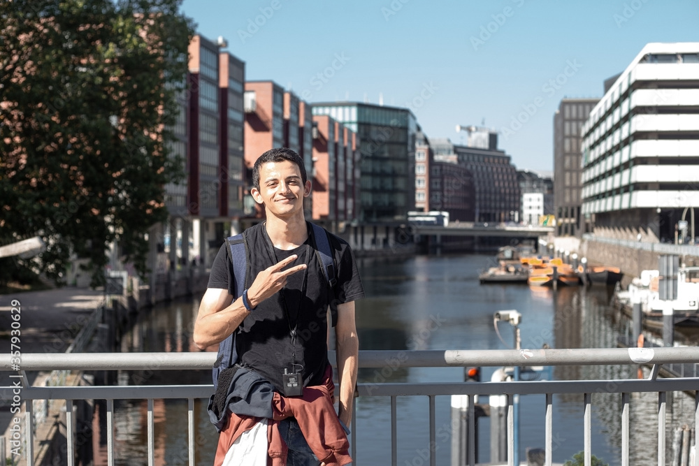 Fototapeta premium Young middle eastern man with backpack walks on eurotrip excursion in downtown of Hamburg in Germany using audio guide and is photographed on background of city canals in sunny summer day