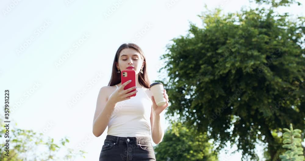 Young Girl is laughing and reading something on her Smartphone while holding a cup of Coffee in other hand. Cheerful young woman communicating on smartphone while walking in park Young Girl is laughing and reading something on her Smartphone while holding a cup of Coffee in other hand. Cheerful young woman communicating on smartphone while walking in park