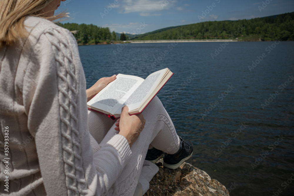 Woman reading a book near to lake in a sweater on a sunny day in nature. A girl sitting on a large rock on the shore of the lake with a book on legs. Relaxation concept. Close up, selective focus