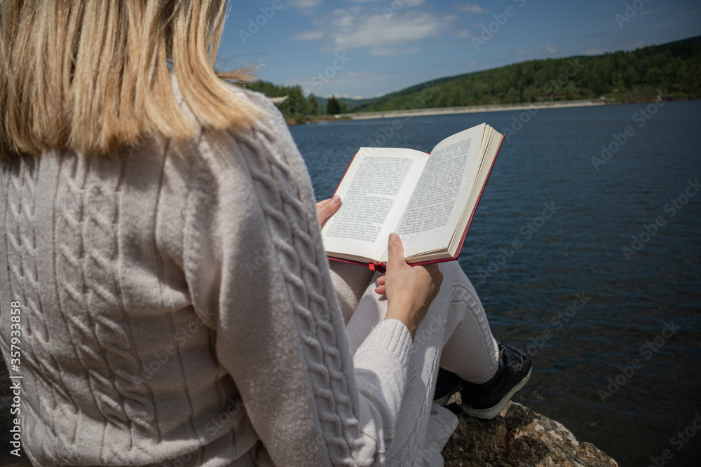 Woman reading a book near to lake in a sweater on sunny day in nature. Blonde girl sitting on a large rock on the shore of the lake with a book on legs. Relaxation concept. Close up, selective focus