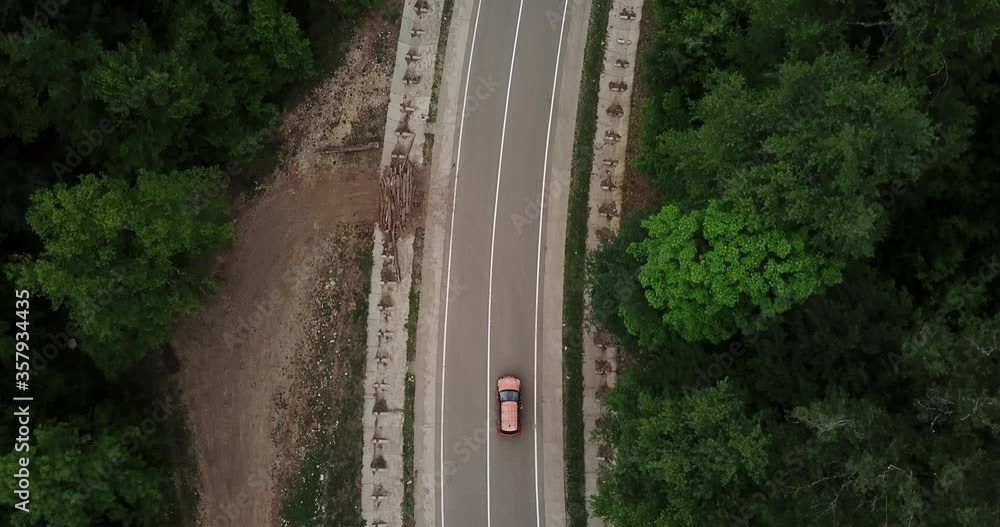 Drone point of view, tracking mode: aerial view flying over two lane countryside forest road with orange car moving green trees of dense woods growing both sides. Car driving along the forest road. Drone point of view, tracking mode: aerial view flying over two lane countryside forest road with orange car moving green trees of dense woods growing both sides. Car driving along the forest road.