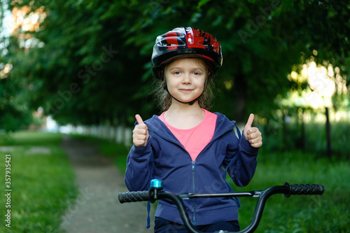 Little girl learns to ride a bike in the park near the home. Kid shows the thumbs up on bicycle. Happy smiling child in helmet riding a cycling.