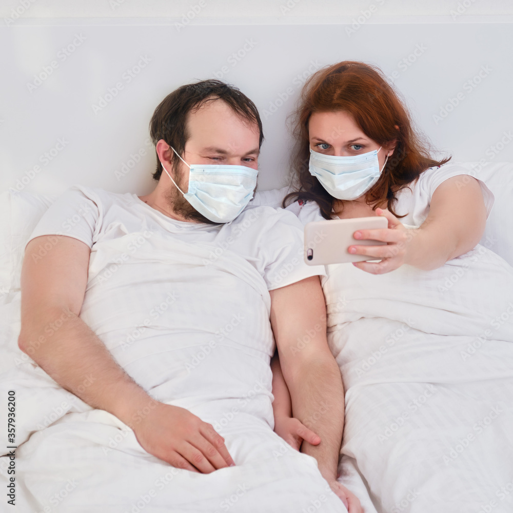 Man and woman look at the phone lying in bed in a medical mask and take a selfie photo. Husband and wife together in isolation due to the virus epidemic.