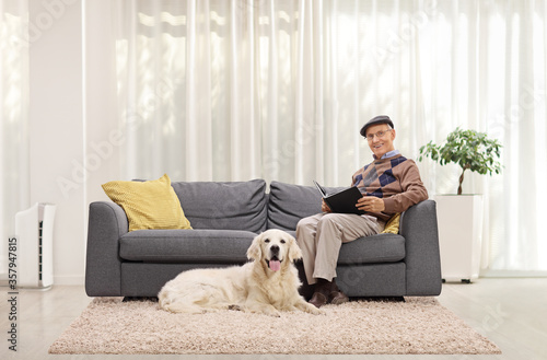 Elderly man at home reading a book on a sofa and his pet dog sitting beside