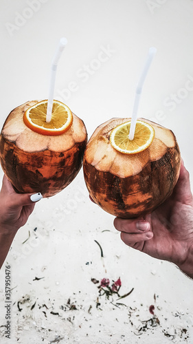 Man and woman holding coconut juice punch cocktails in hands. Close up view with white sand in the background. Serene island lifestyle. Remote destination for joyful and happy honeymoon vacation.