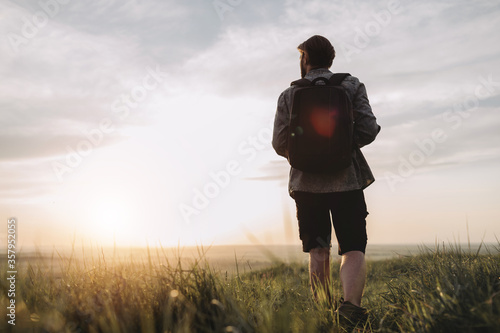 Back view of mature man in denim shirt and black shorts walking on high hill with big backpack. Travel photographer discovering new place for shooting nature.