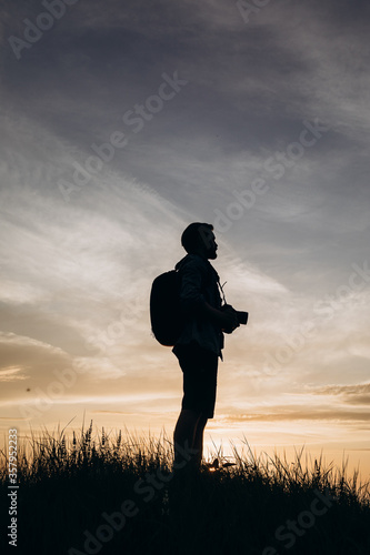 Mature man in silhouette with backpack and digital camera standing outdoors with background of amazing sunset. Concept of professional photography and travelling.