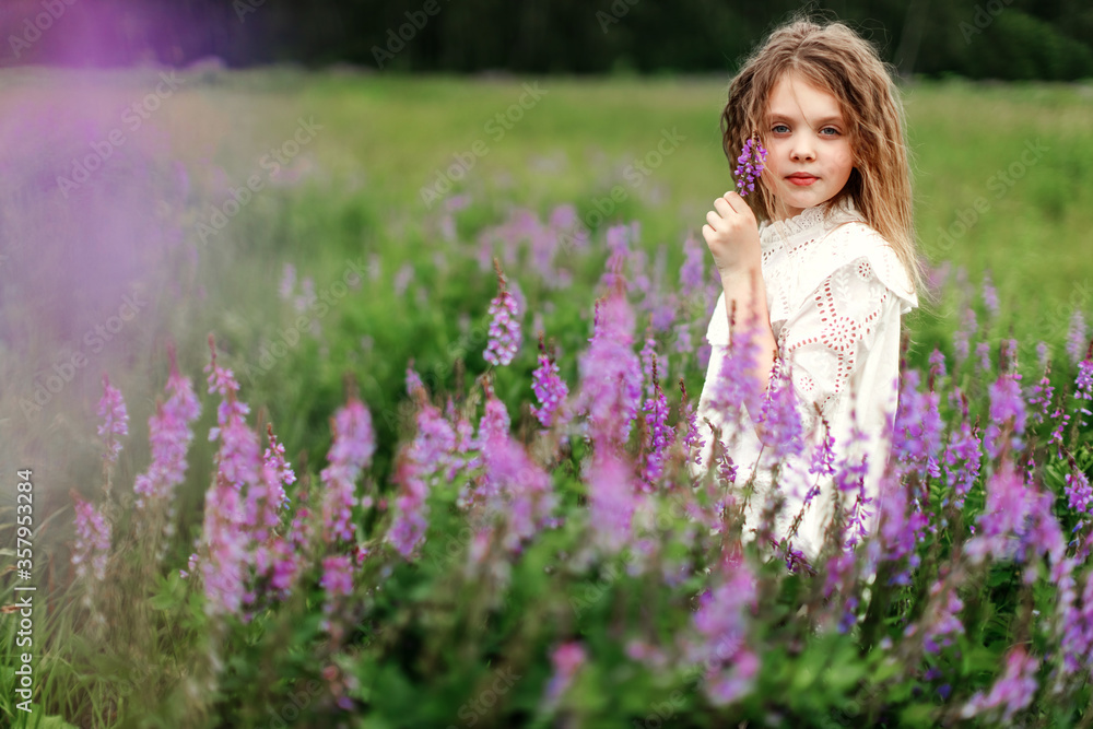 A little girl looks at camera in a white dress with beautiful flowers in the field in summer. Concept of happy childhood. Copy space for text A little girl looks at camera in a white dress with beautiful flowers in the field in summer. Concept of happy childhood. Copy space for text