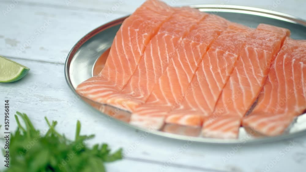 Close-up slow-motion of sliced fresh raw red salmon.
Raw salmon in a silver plate on the wooden table with lemon and herbs/greens ingredients on the side. Concept of healthy seafood. Salmon in 4K.