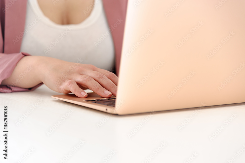 Naklejka premium Woman wearing a pink jacket and using a gold laptop, incredibly thin, light and perfectly portable notebook open on a white wooden table.