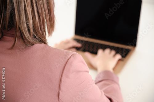Redhead woman wearing a pink jacket and typing on the keyboard using a gold laptop, incredibly thin, light and perfectly portable notebook open with blank black screen on a white wooden table.