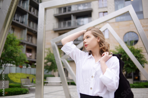 Student, manager, realtor, girl in a white shirt on the background of the building posing. Beautiful blonde with short hair. Model with red lips and a black jacket. © MoreThanProd