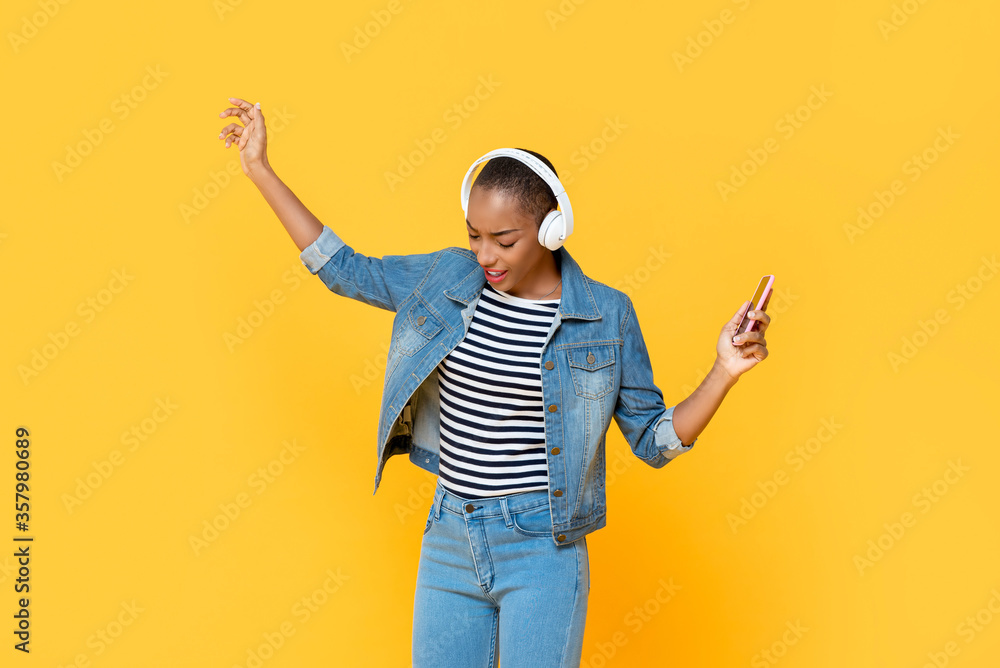 Happy African American woman wearing headphones listening to music online and dancing on isolated yellow background