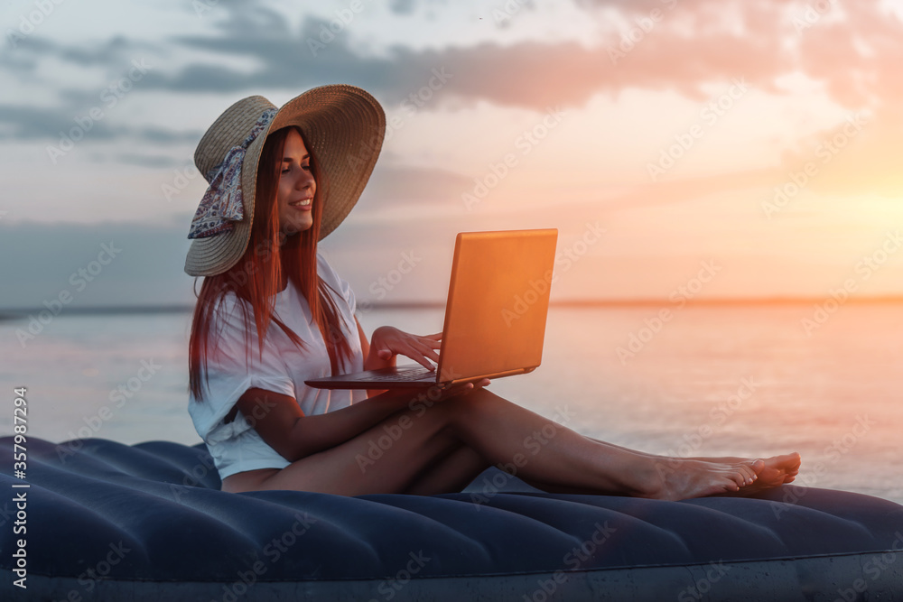 Young woman in straw hat working on a laptop on the beach.  Working on vacation concept.