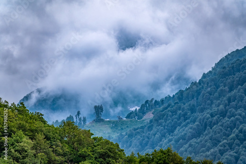 High mountain with green slopes hidden in clouds and fog. Green mountain slope with cable cars and ski runs at overcast summer day.