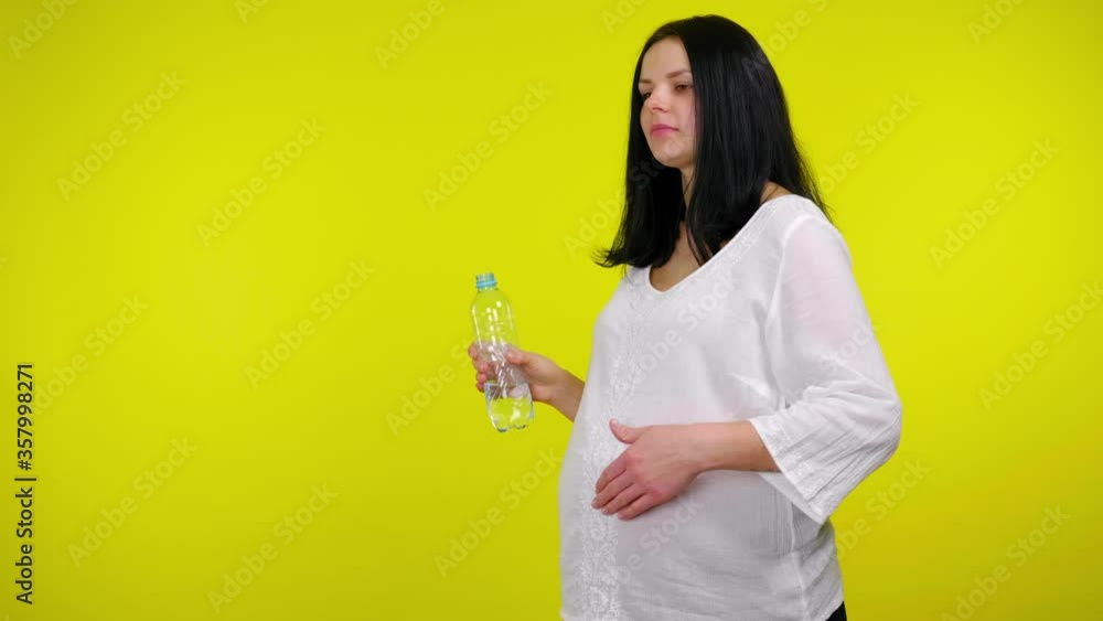 Young pregnant woman in a white blouse stands with water in a plastic bottle and massages stomach with baby inside on a yellow background with copy space. 4K slow motion footage Young pregnant woman in a white blouse stands with water in a plastic bottle and massages stomach with baby inside on a yellow background with copy space. 4K slow motion footage