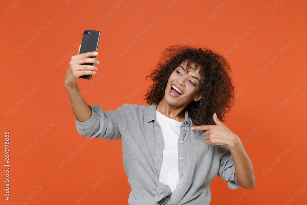 Excited young african american woman in casual clothes isolated on orange background. People lifestyle concept. Mock up copy space. Doing selfie shot on mobile phone, pointing index finger on herself. Excited young african american woman in casual clothes isolated on orange background. People lifestyle concept. Mock up copy space. Doing selfie shot on mobile phone, pointing index finger on herself.