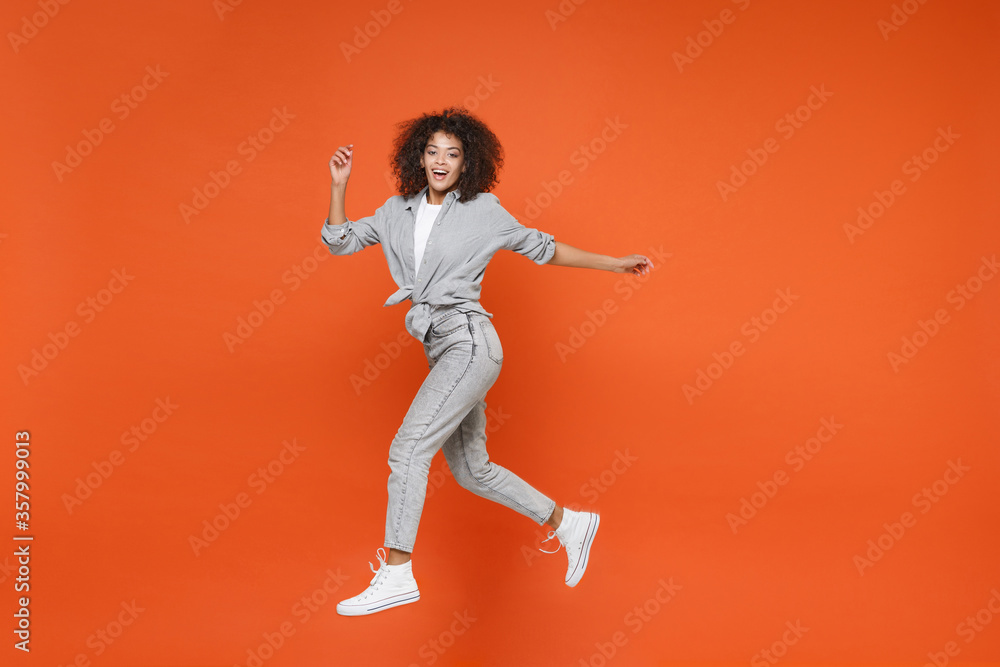 Cheerful young african american woman girl in gray casual clothes posing isolated on orange background studio portrait. People lifestyle concept. Mock up copy space. Jumping, spreading hands and legs.