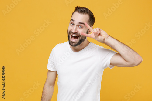 Excited young bearded man guy in white casual t-shirt posing isolated on yellow wall background studio portrait. People sincere emotions lifestyle concept. Mock up copy space. Showing victory sign.