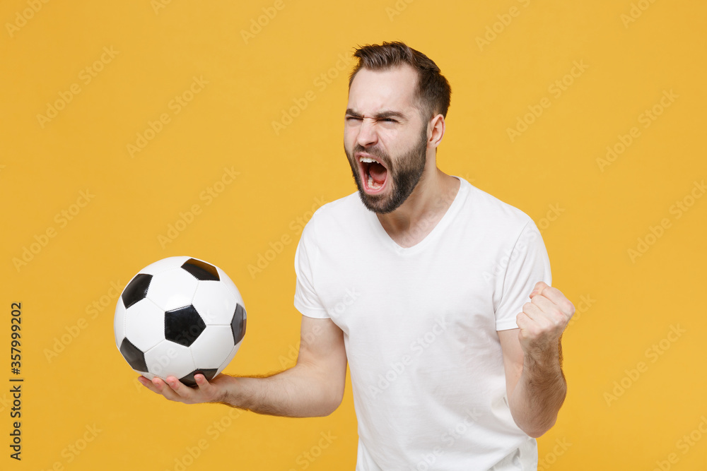 Angry young bearded guy football fan in white t-shirt isolated on yellow wall background. People sport family leisure concept. Cheer up support favorite team with soccer ball screaming clenching fist. Angry young bearded guy football fan in white t-shirt isolated on yellow wall background. People sport family leisure concept. Cheer up support favorite team with soccer ball screaming clenching fist.