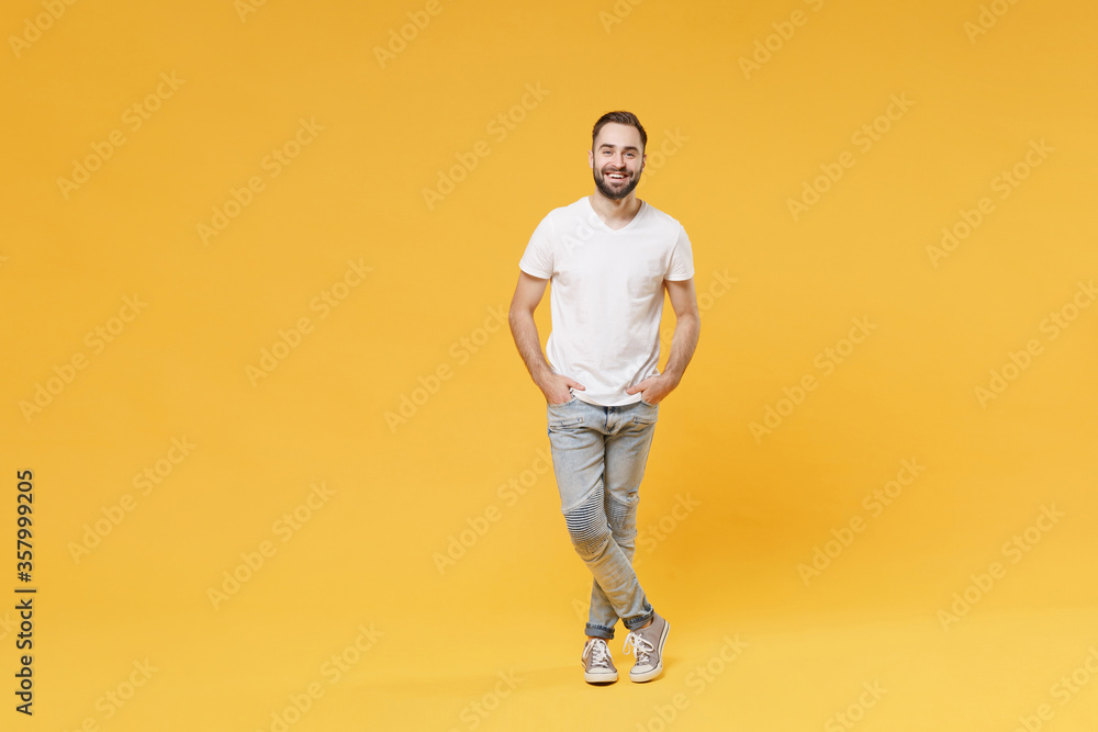 Fototapeta premium Smiling young bearded man guy in white casual t-shirt posing isolated on yellow background studio portrait. People sincere emotions lifestyle concept. Mock up copy space. Holding hands in pockets.