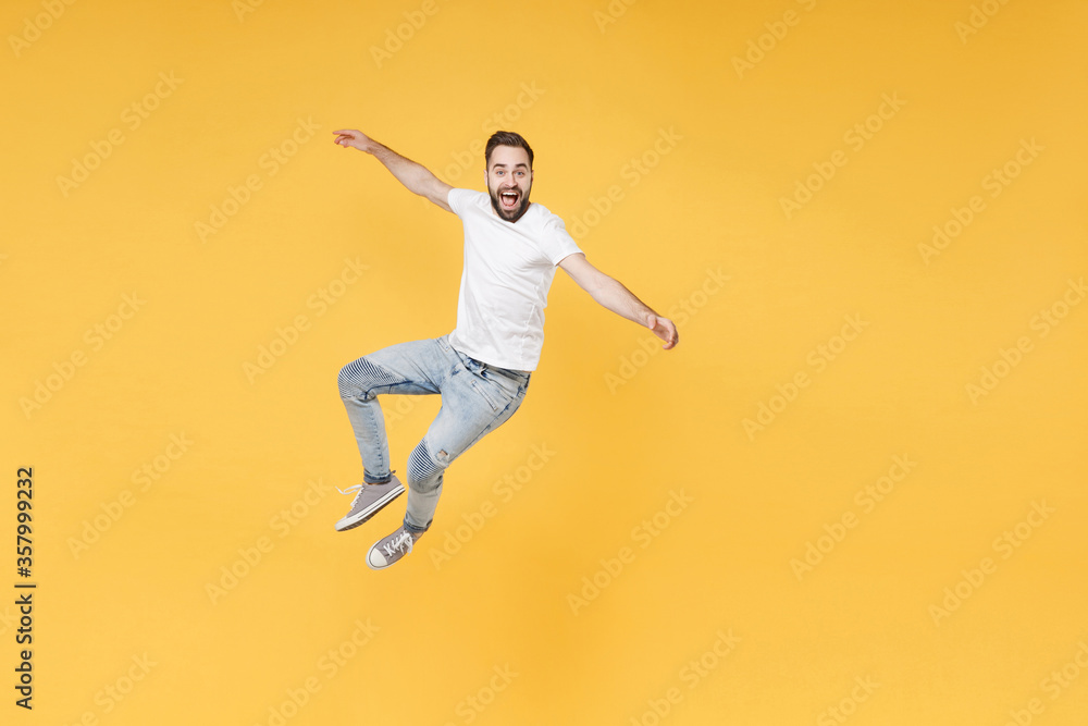 Excited young bearded man guy in white casual t-shirt posing isolated on yellow wall background studio portrait. People sincere emotions lifestyle concept. Mock up copy space. Jumping spreading hands.